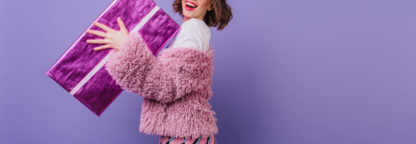 Laughing carefree woman holding sparkle present box on purple background. Indoor photo of stunning birthday girl with wavy hair posing with gift.