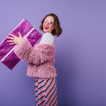 Laughing carefree woman holding sparkle present box on purple background. Indoor photo of stunning birthday girl with wavy hair posing with gift.